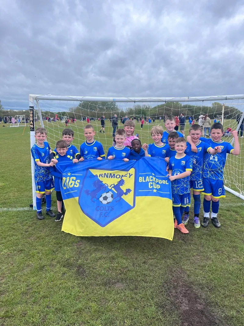 A squad holding the Blackpool Cup flag in a goal on a grass pitch