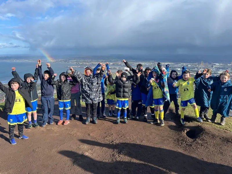 Children on top of Cave Hill with their arms up and a rainbow over the lough behind