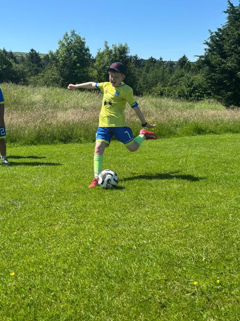 A child in Colts kit taking a full swing at a football on a foot golf course