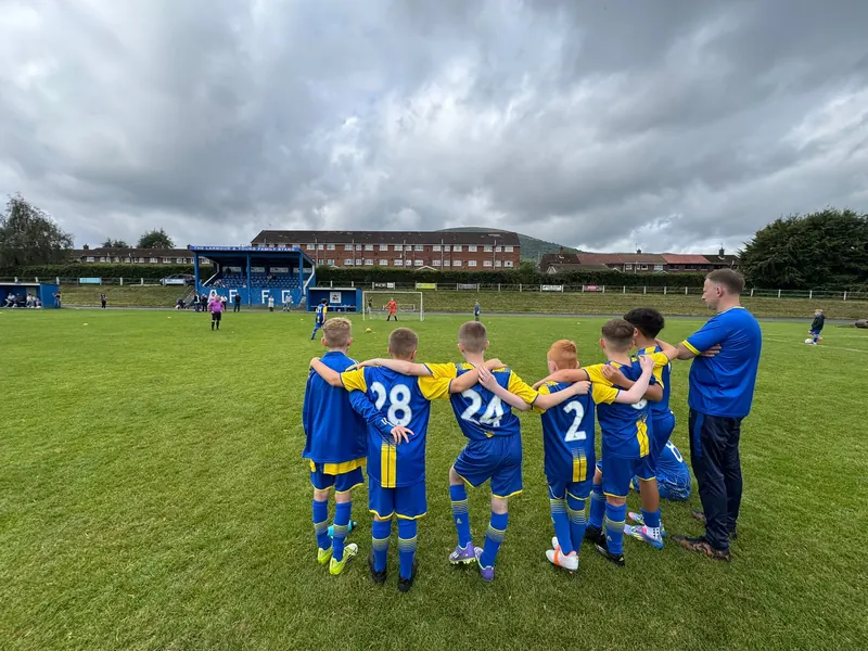 A team huddle seen from behind, arms round each other's shoulders, a coach beside them